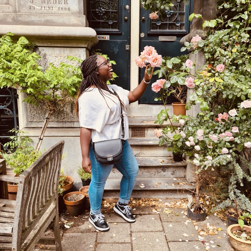 Tatiana lacht terwijl ze een roze roos vasthoudt bij een gevel vol bloemen.